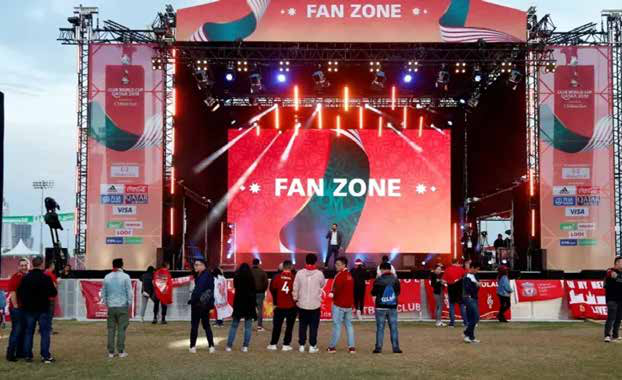 Outdoor fan zone stage with a large LED screen and spectators on the grass